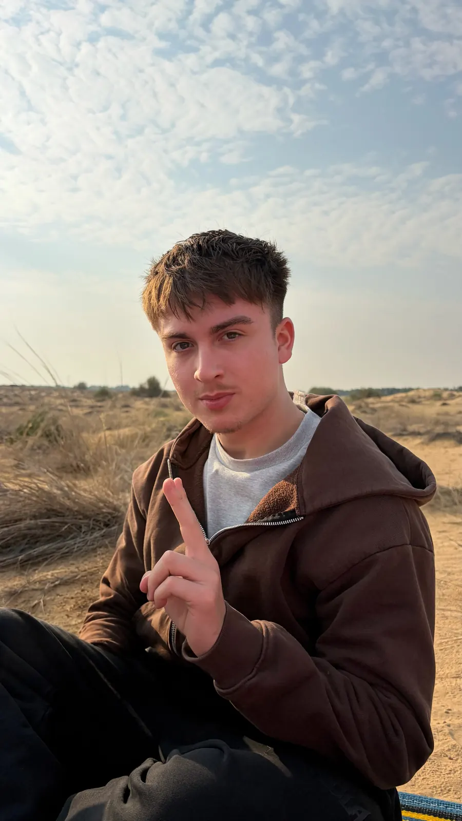 Rafa Villaseñor sitting outdoors in a brown hooded jacket, dunes and open sky behind him.